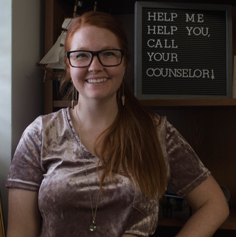 Lindsey Edwards poses in her office with a sign that says "help me help you, call your counselor."