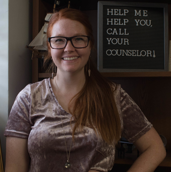 Lindsey Edwards poses in her office with a sign that says help me help you, call your counselor.