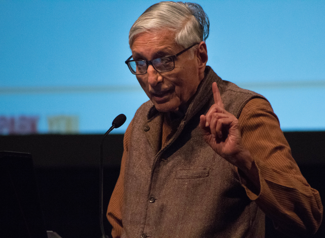 Rajmohan Gandhi speaking on stage. He holds up his pointer finger to emphasize his point.