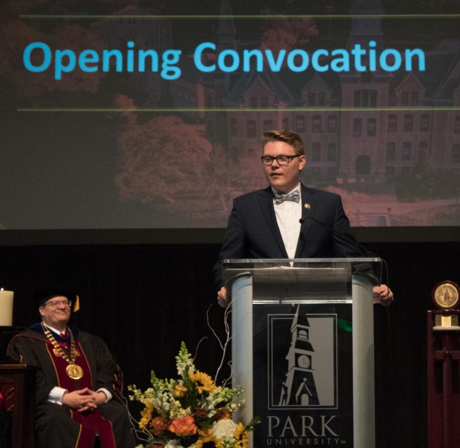 Christian Leonard stands on stage in a suit to give a speech. In the background Park University president Greg Gunderson smiles as he watches.